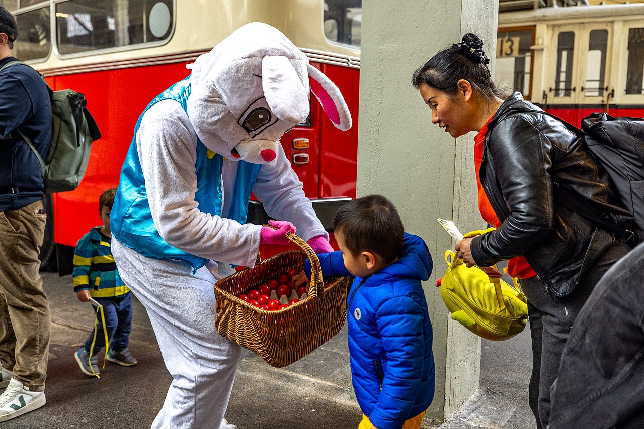 Osterhase hält einem Kind einen Korb voller Ostereier hin und das Kind greift in den Korb und sucht sich eines aus