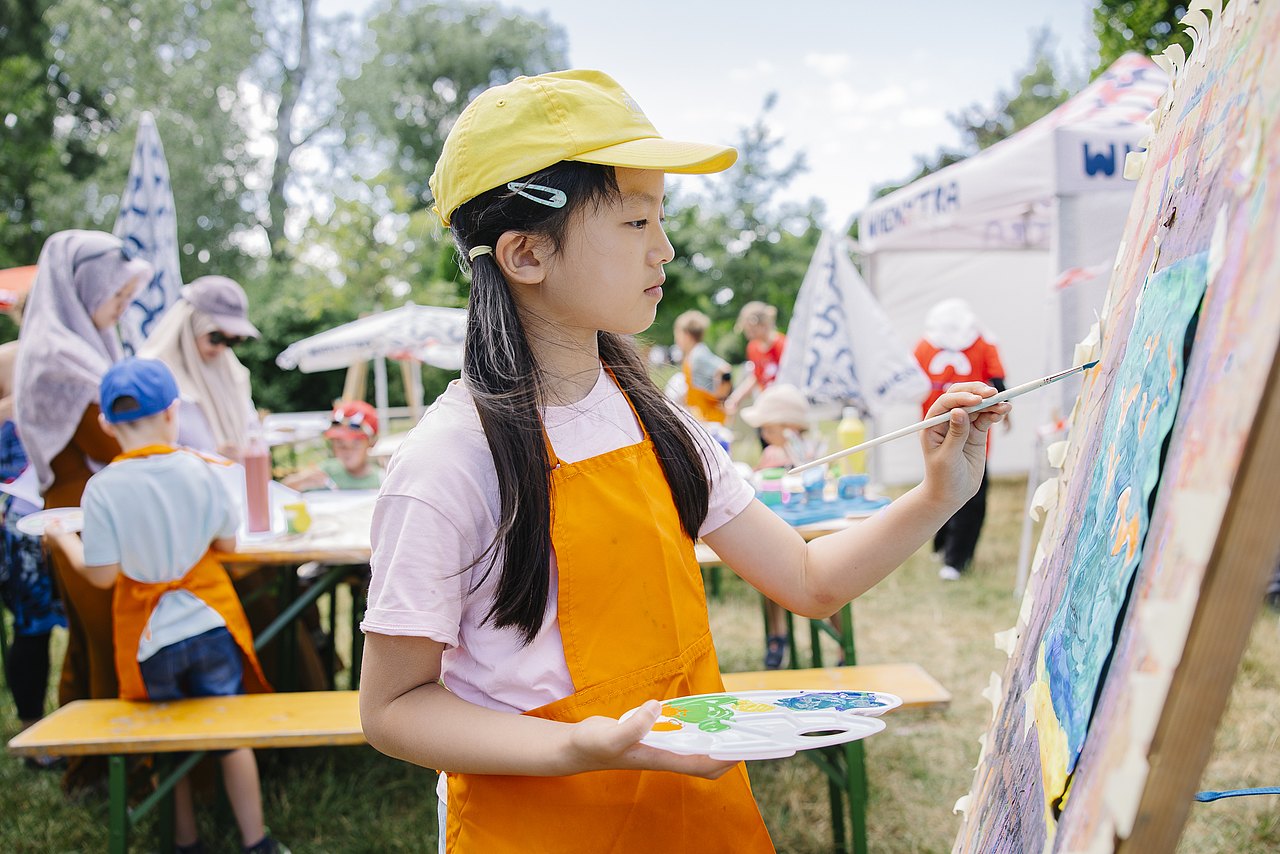 Kind mit oranger Schürze und gelber Kappe malt an einer Staffelei mit einem Pinsel ein buntes Bild. In der Hand hält es eine Farbpalette. Im Hintergrund sieht man, dass es bei einer Veranstaltung in einem Park ist: mehrere Personen, Heurigentische und Bänke, Sonnenschirme, Wiese und Bäume
