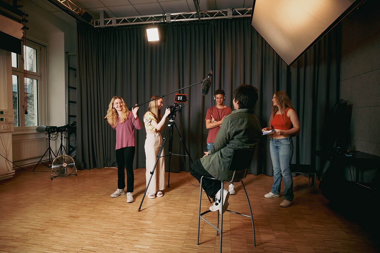 Sechs Jugendliche arbeiten in einem Studio an einer Videoproduktion: Eine Jugendliche hält eine Tonangel, eine weitere bedient die Kamera auf einem Stativ, ein Jugendlicher sitzt auf einem Stuhl und wird interviewt, während drei weitere Jugendliche bei der Produktion assistieren.