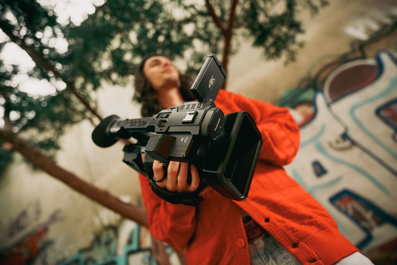 Ein Jugendlicher steht im Freien vor einer Wand mit Graffiti und hält eine Panasonic-Videokamera in den Händen.