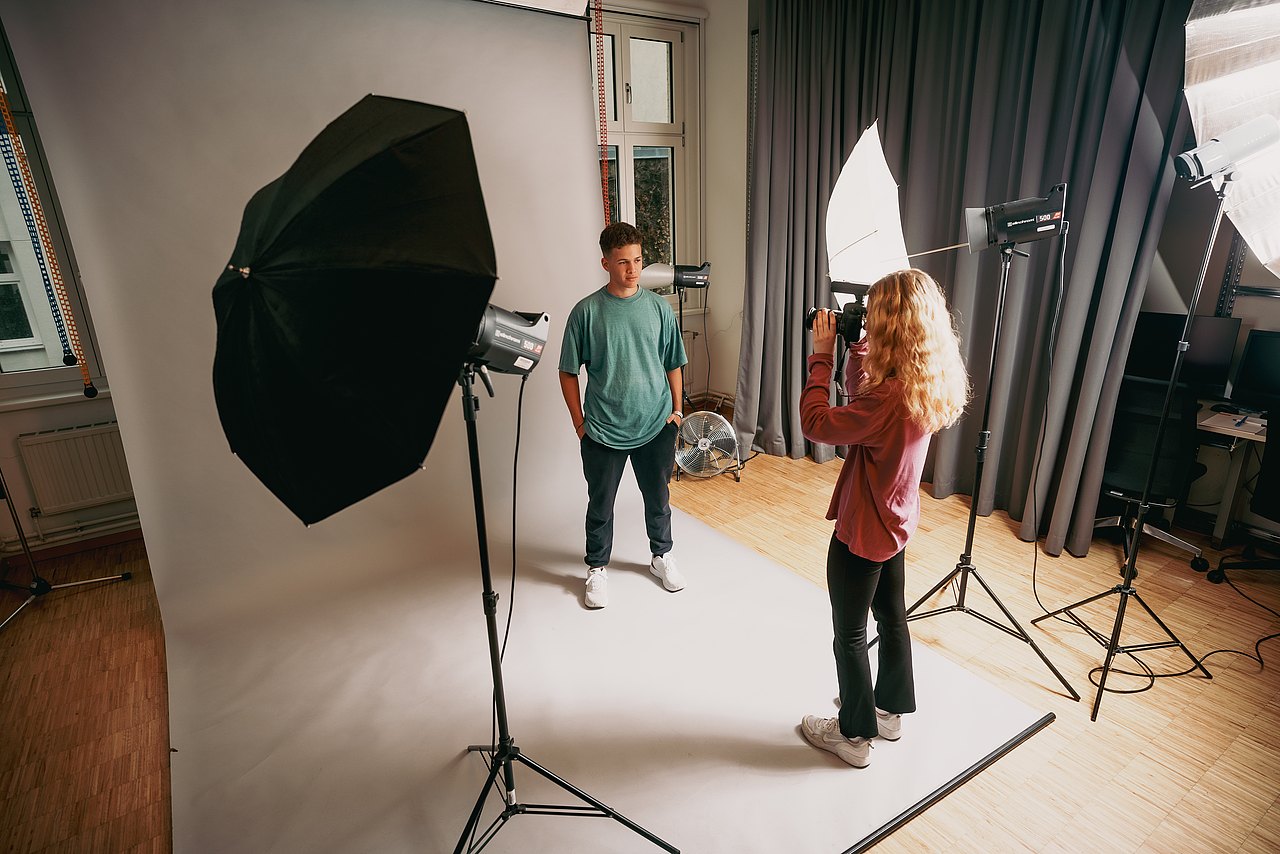 Eine Jugendliche fotografiert einen Jugendlichen vor einem weißem Hintergrund in einem Fotostudio mit professioneller Beleuchtung.