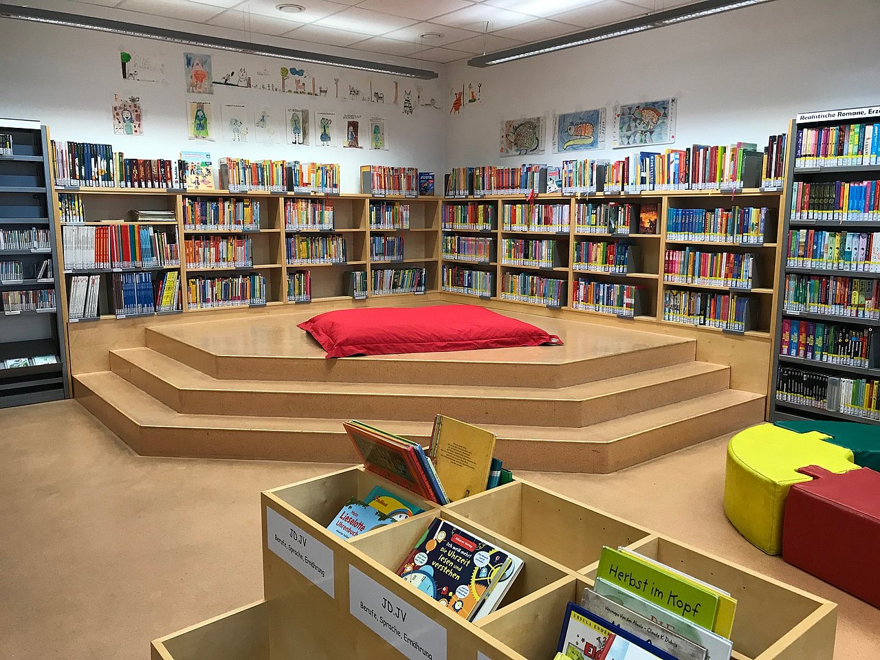 Ein Holzpodium mit zwei Bücherwänden, auf dem ein langer roter Sitzpolster liegt. An der Wand hängen Kinderzeichnungen, vor den Stiegen des Podiums stehen Bücherkisten.