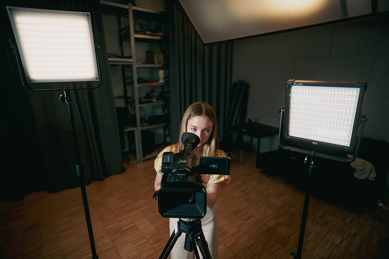 Eine Jugendliche steht zwischen zwei LED-Flächenleuchten und schaut durch den Sucher einer Panasonic-Videokamera.