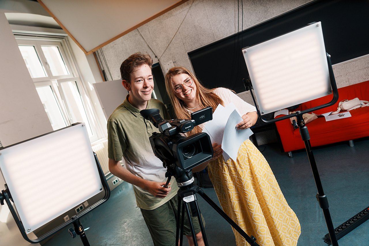 Zwei Jugendliche stehen in einem Studio hinter einer Videokamera und zwei Videoleuchten.