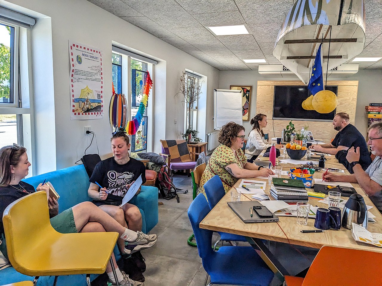 6 Personen sitzen an einem Tisch und auf einem Sofa und führen jeweils zu zweit Gespräche miteinander.