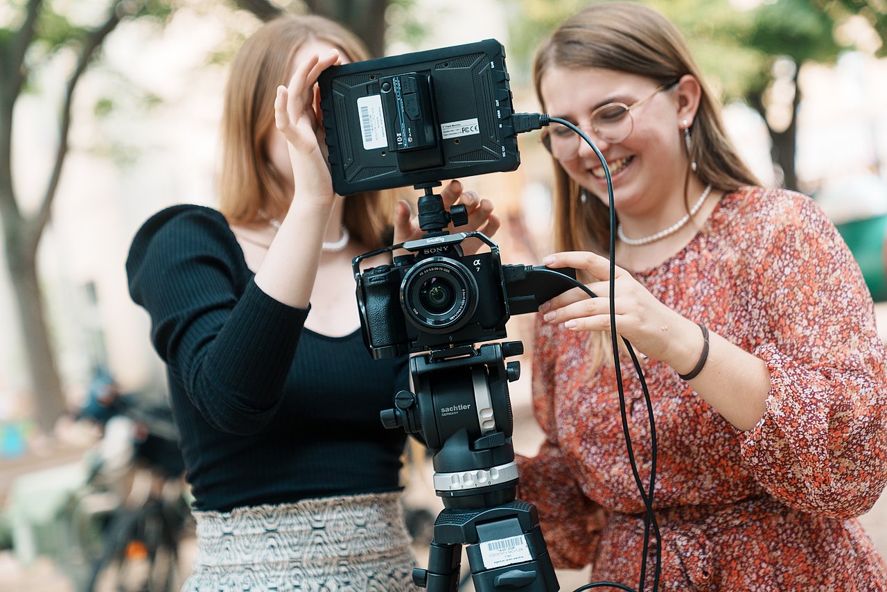 Zwei Jugendliche arbeiten im Freien mit einer professionellen Sony-Kamera auf einem Stativ mit externem Monitor und richten gemeinsam die Kameraeinstellungen ein.