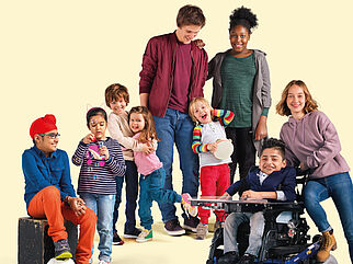 Eine Gruppe junger Menschen im Alter zwischen 5 und 20 Jahren. Ein Kind sitzt m Rollstuhl, ein anderes sitzt auf einer Kiste, sein Fuß ruht auf einem Fußball. Alle lächeln. Ein Mädchen bläst Seifenblasen.  