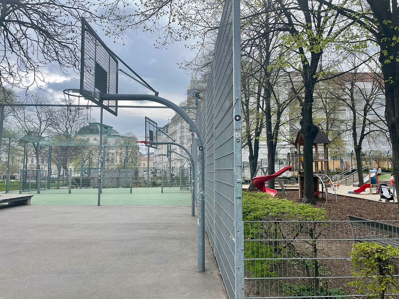 Ballspielkäfig am Spielplatz Weghuberpark Auf der linken Seite sieht man die Ballspielkäfige mit Fußballtoren und Baskettballkörben, auf der rechten Seite sieht man im Park drinnen die Kleinkinderspielgeräte