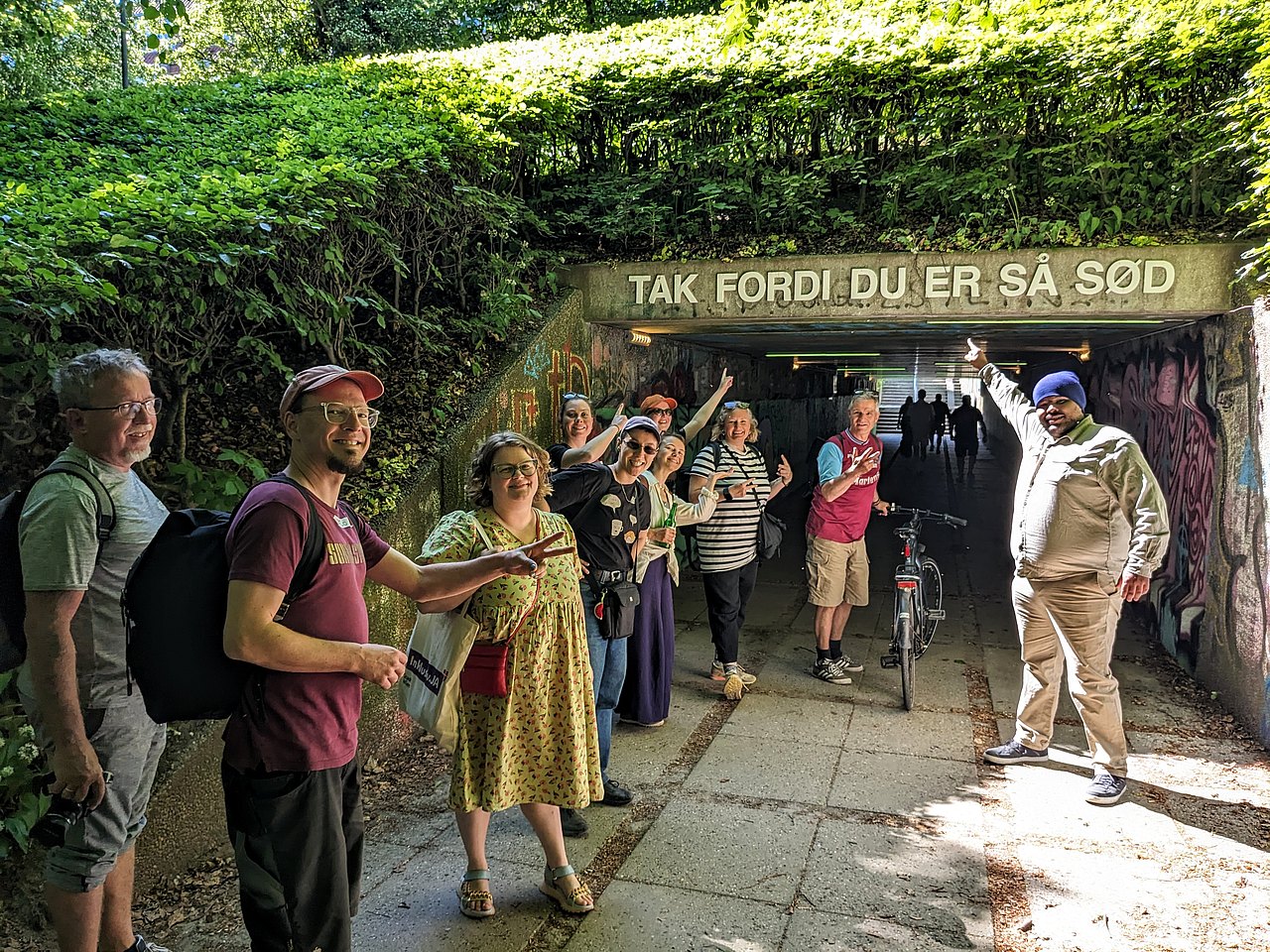 Eine Gruppe von Personen verschiedenen Alters mit bunter Kleidung steht vor einer Unterführung und zeigt auf einen Schriftzug, wo auf Dänisch steht "Danke dass du so süß bist"