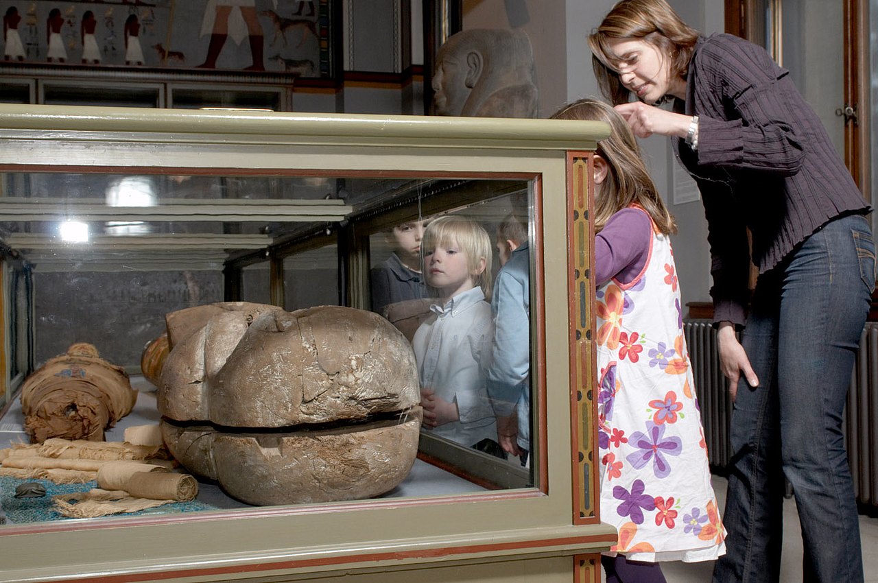 Kinder und eine Erwachsene in der Ägyptisch-Orientalischen Sammlung des Kunsthistorischen Museums. Das Bild zeigt eine Vitrine mit einem Sarkophag. Kinder und eine Erwachsene schauen hinein.