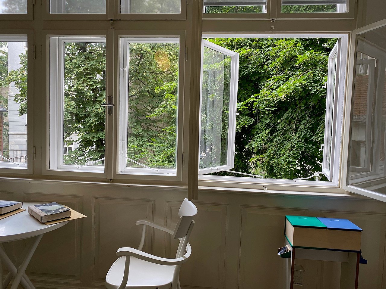 Blick in den grünen Hof weißer Tisch udn Sessel im Hintergrund großes Kastenfenster mit Blick in den grünen Hof