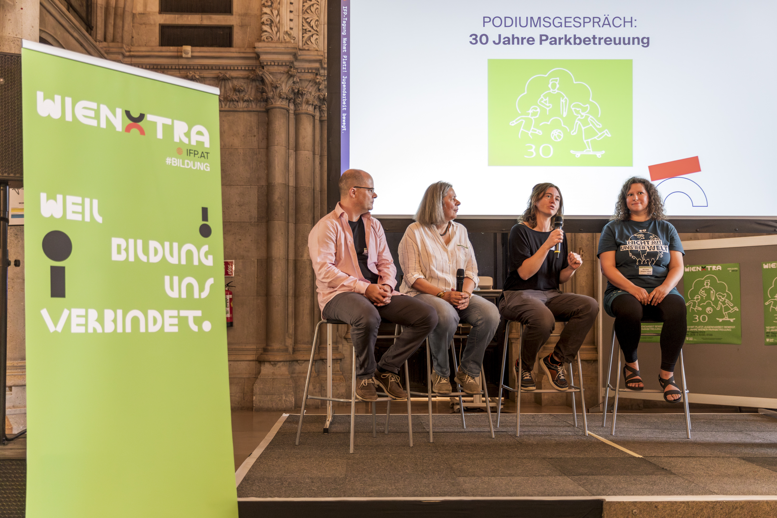 Vier Personen sitzen auf Hochstühlen auf einer Bühne und schauen sich an. Im Hintergrund ist eine Leinwand mit Projektion zu sehen. Auf der linken Seite steht ein Aufsteller.