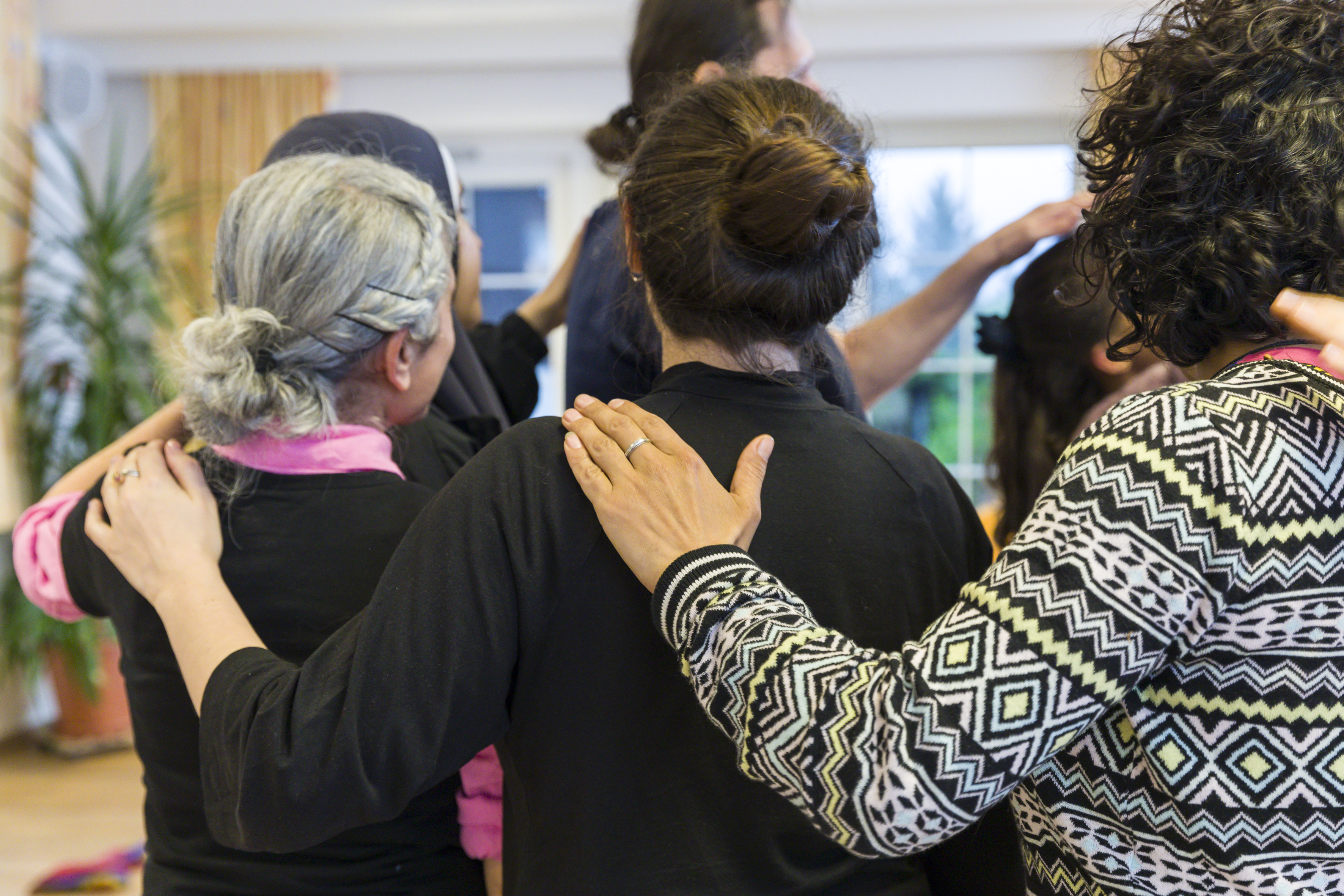 Mehrere Personen stehen in einem Kreis. Die Personen wenden einander den Rücken zu. Jede Person hat ihre linke Hand auf die Schulter der vorderen Person gelegt.