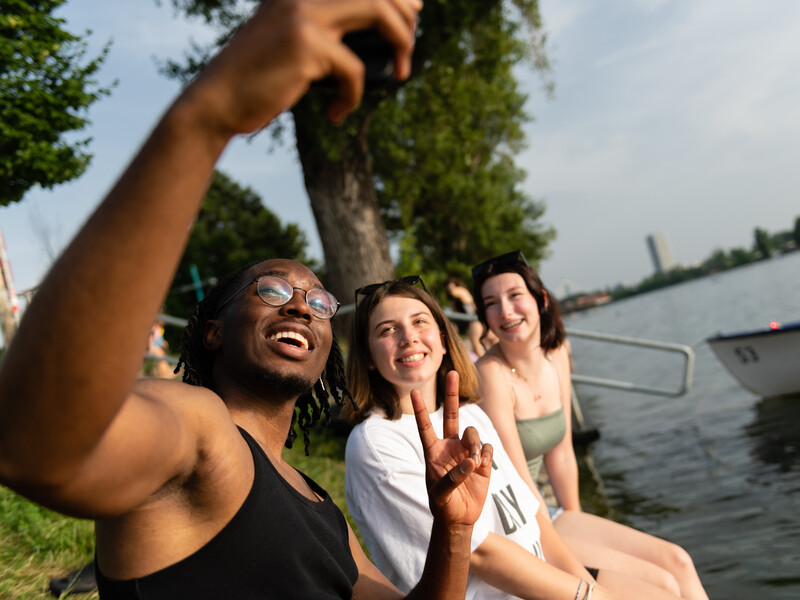 Drei Jugendliche sitzen am Wasser und machen ein Selfie.