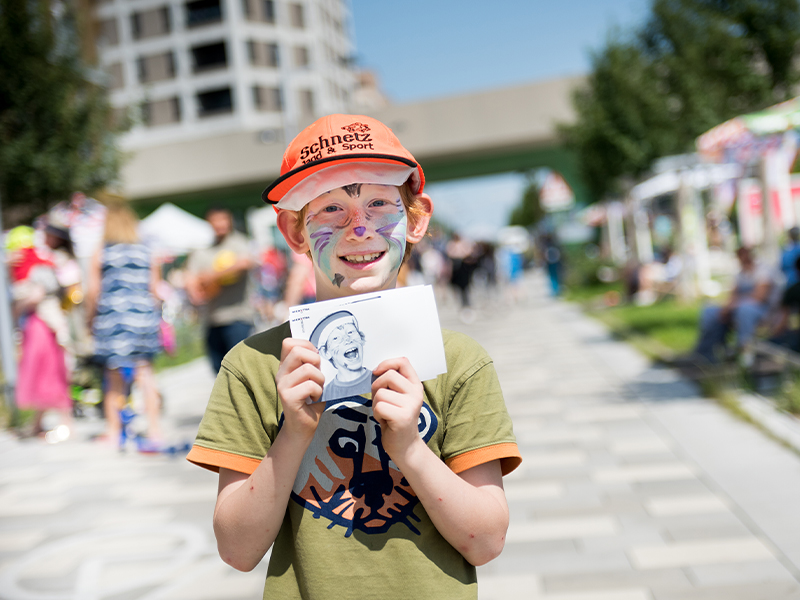 Ein Junge mit oranger Kappe und Gesichtsbemalung hält ein Foto hoch.